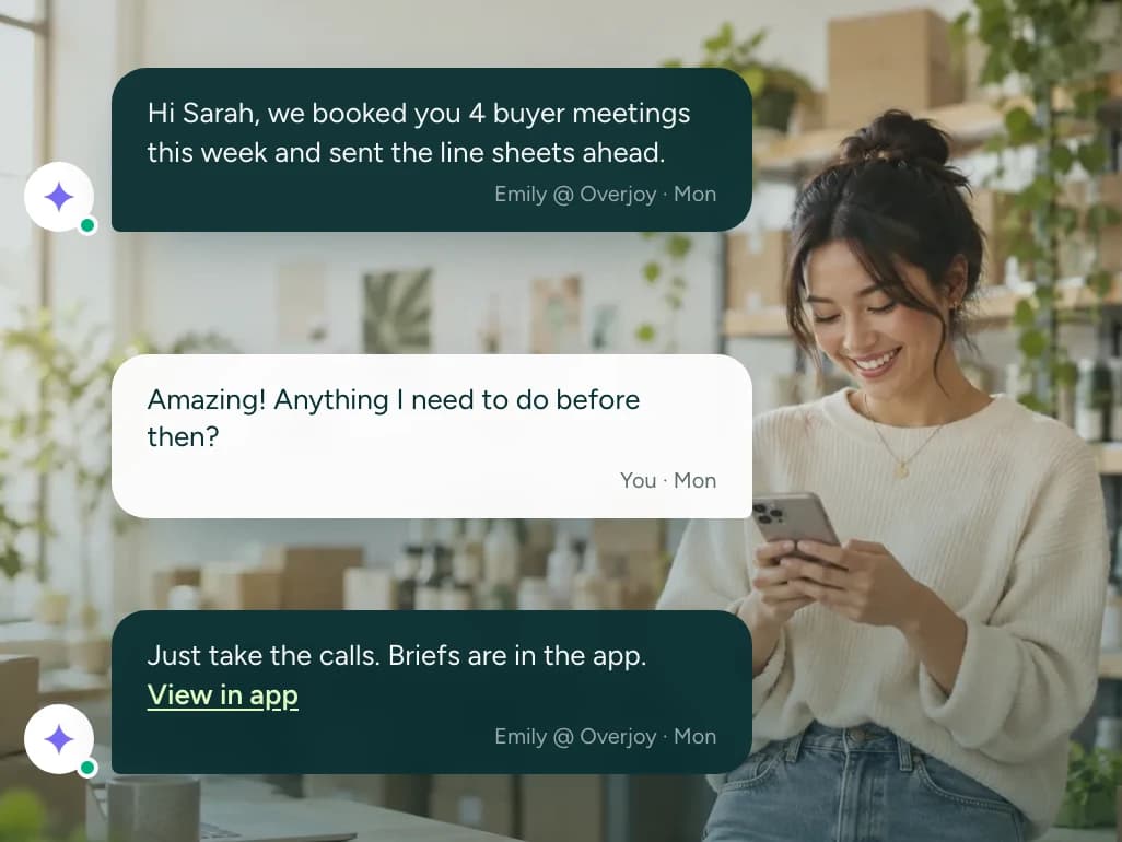 A brand founder smiling at her phone, over a conversation with her Overjoy specialist: four buyer meetings booked this week with the line sheets sent ahead, and the briefs waiting in the app
